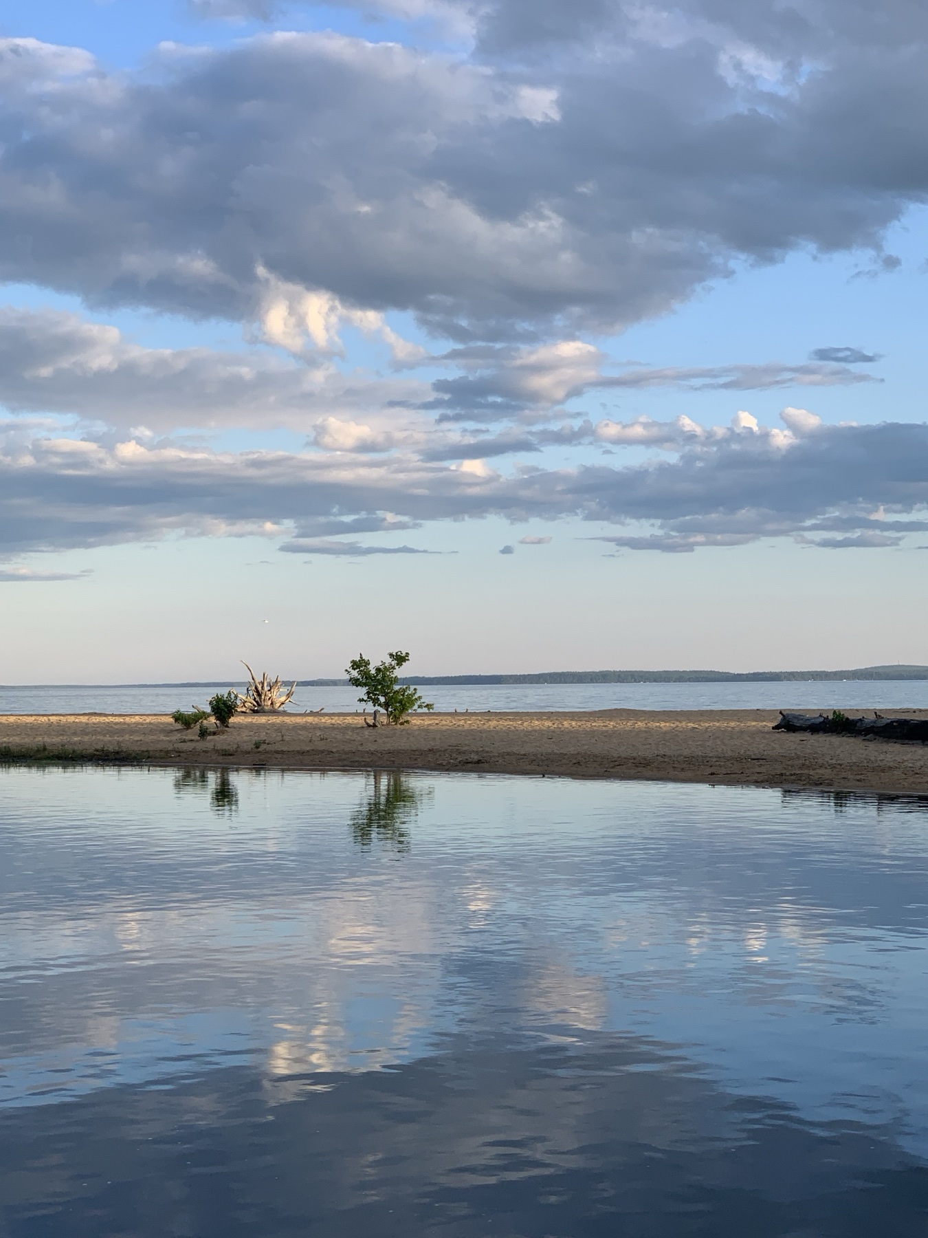 A sandy shoreline between the Songo River and the open water of Sebago Lake beneath a broad cloud-filled sky.