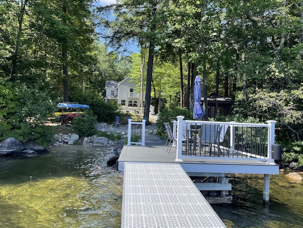 A wooden deck extending over a clear stream, surrounded by lush greenery and a building in the background.