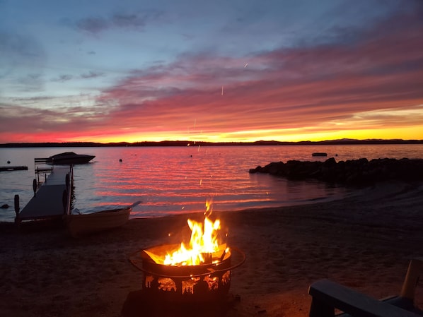 A beach scene with a fire pit in the foreground and boats on the water.
