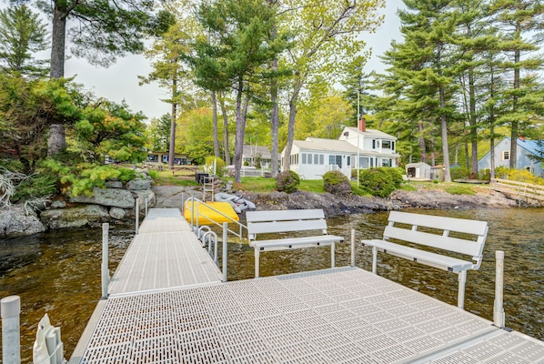 A wooden dock leading to a house by a lake, with benches and a small shed.