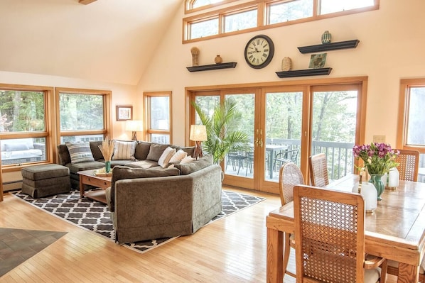A spacious living room with a sectional sofa, a dining table with chairs, and large windows offering a view of the outdoors.