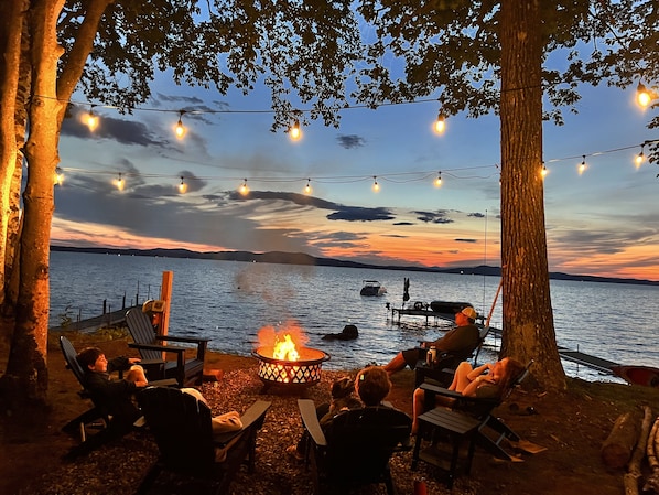 An outdoor seating area with chairs and a fire pit, overlooking a body of water with a sunset in the background.