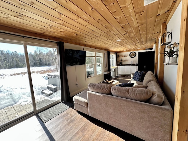 A cozy cabin interior with a wooden ceiling, a large sliding glass door to a snowy outdoor area, a kitchenette, and a comfortable seating area.