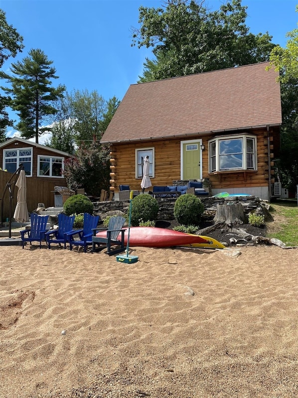 A log cabin with a sandy yard, blue Adirondack chairs, a red canoe, and a stone wall.