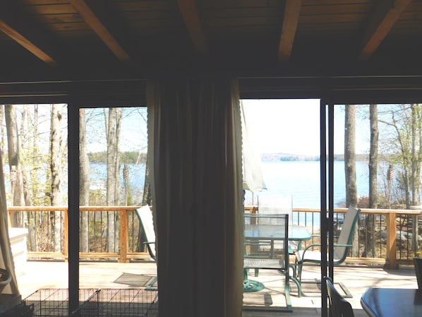 A view from a room with large windows overlooking a lake, with outdoor furniture and a wooden deck.