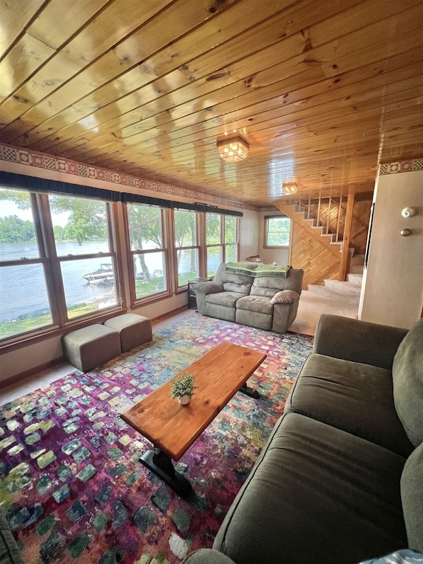 A cozy cabin interior with a wooden ceiling, large windows overlooking a lake, a sofa, a coffee table, and a staircase leading to an upper level.