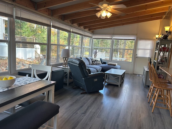 A spacious living area with a large window, wooden ceiling, and a ceiling fan.